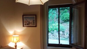 Une chambre à coucher avec un lit, une lampe, une fenêtre donnant sur un paysage verdoyant et un tableau accroché au mur.