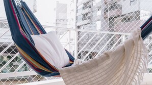 Una silla hamaca con un cojín blanco en un balcón con barandillas blancas y vistas a edificios.