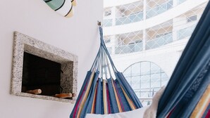 Una silla hamaca en una habitación con pared blanca, una máscara decorativa y una ventana con vistas a un edificio.