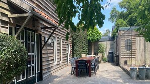 Un bâtiment en bois doté d’un patio comprenant une table et des chaises, entouré de verdure et bordé d’un chemin pavé.