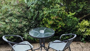 A round glass table with two wicker chairs, surrounded by a garden with dense foliage. - Spacious Apartment with Private Terrace (Newtownards)
