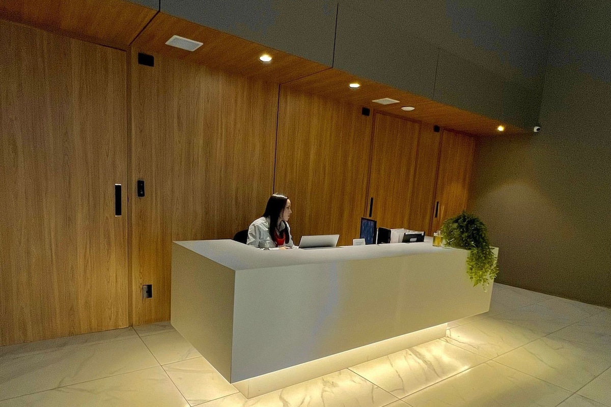 A reception desk with a woman seated behind it, a potted plant to the right, and wooden paneling on the walls.
