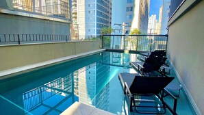 Un área de piscina en la azotea con agua azul cristalina, barandilla de vidrio y sillones de lounge modernos.