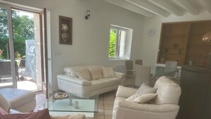 Una sala de estar moderna con sofá, sillón y mesa de centro de vidrio. Hay una puerta corrediza de vidrio que da a un área exterior.