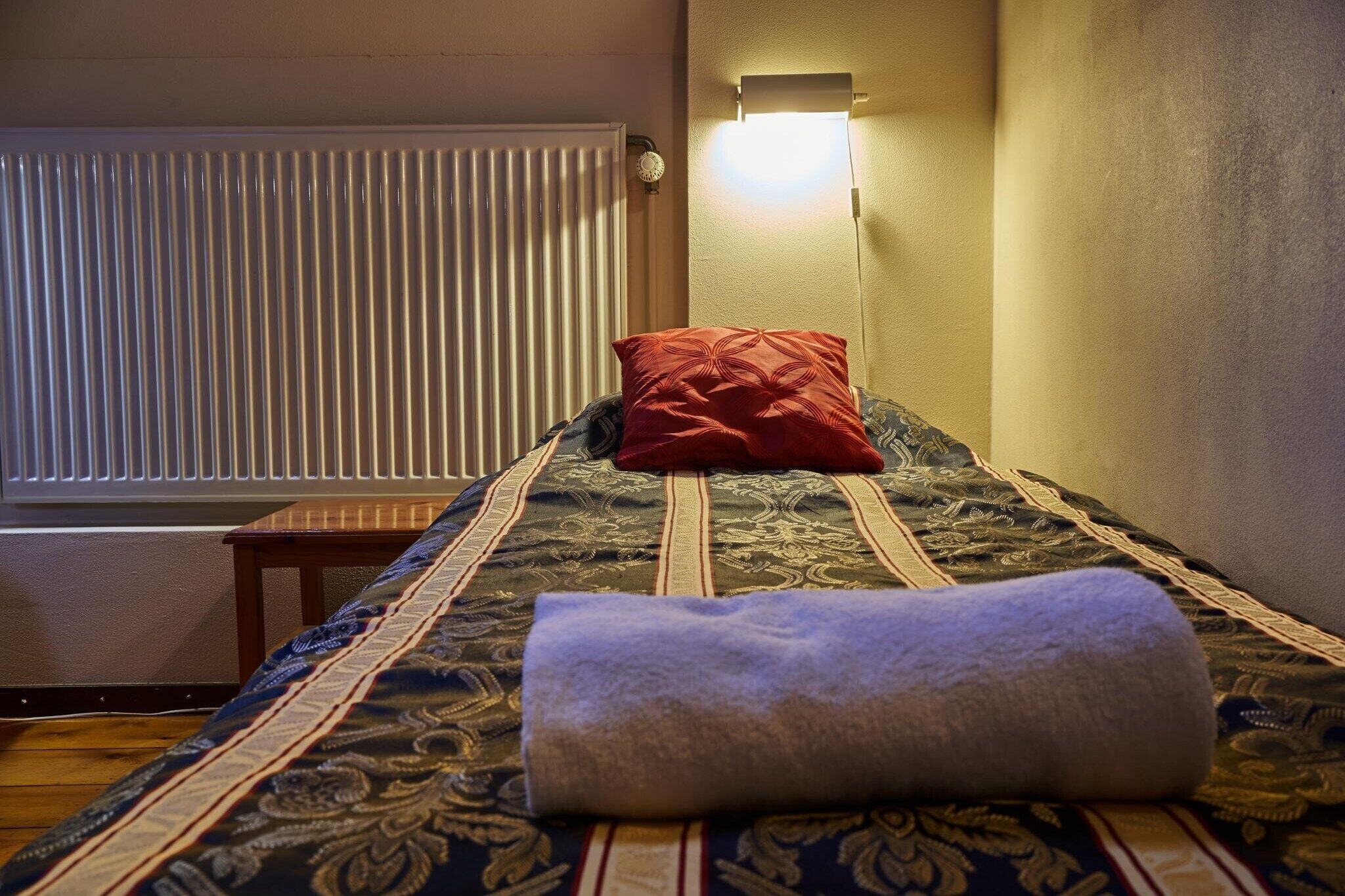 Un lit bien fait, avec une couvre-lit à motifs, une serviette enroulée et un oreiller rouge.