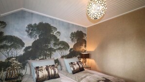Quarto com mural de árvores na parede, cama com roupa de cama estampada, luminária decorativa e luminária pendente.