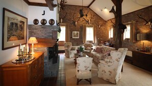 Uma sala de estar rústica com paredes de tijolo aparente, vigas de madeira e decorações de chifres. Há uma lareira, poltronas florais e um armário de madeira com um candelabro.