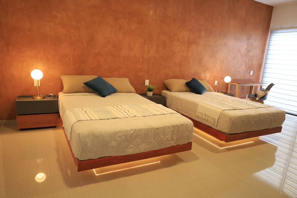 Quarto moderno com duas camas, mesas de cabeceira e uma pequena planta.