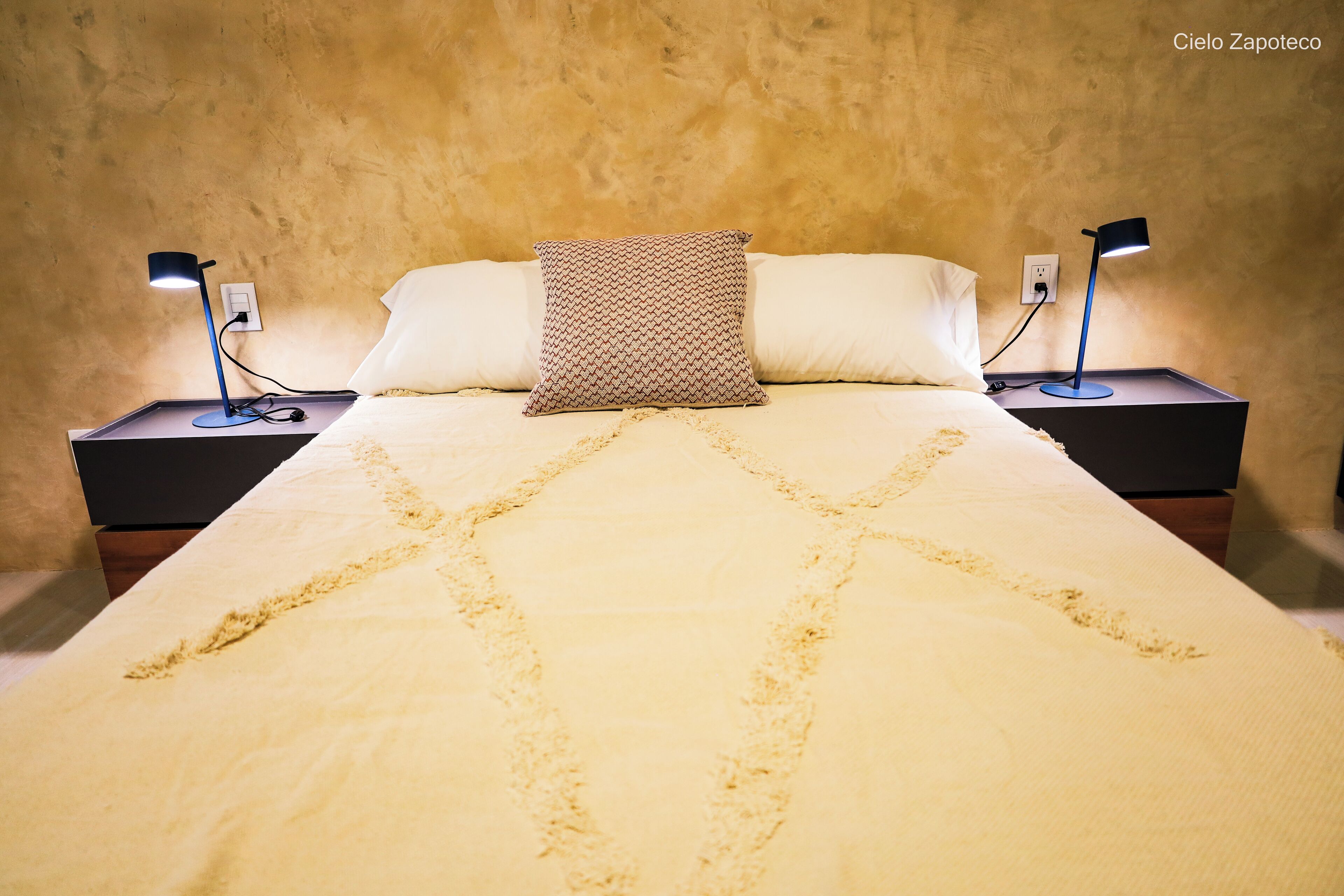 Una cama con un cojín estampado, dos lámparas de noche y una pared con textura.
