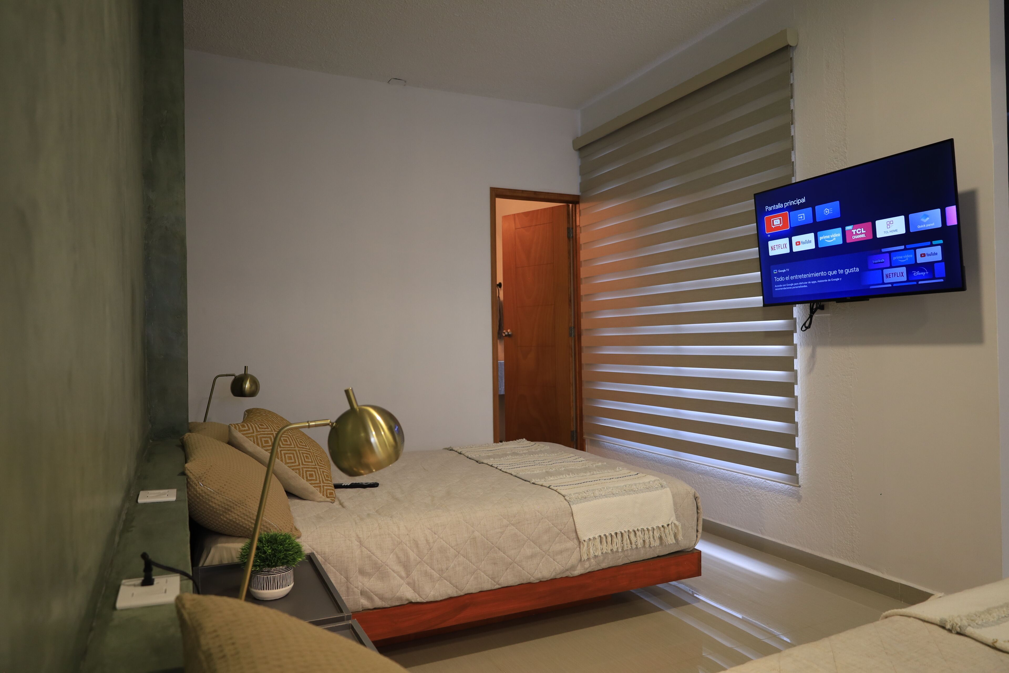 Une chambre à coucher avec un lit, un téléviseur fixé au mur, une table de chevet avec une lampe et une fenêtre avec des stores.