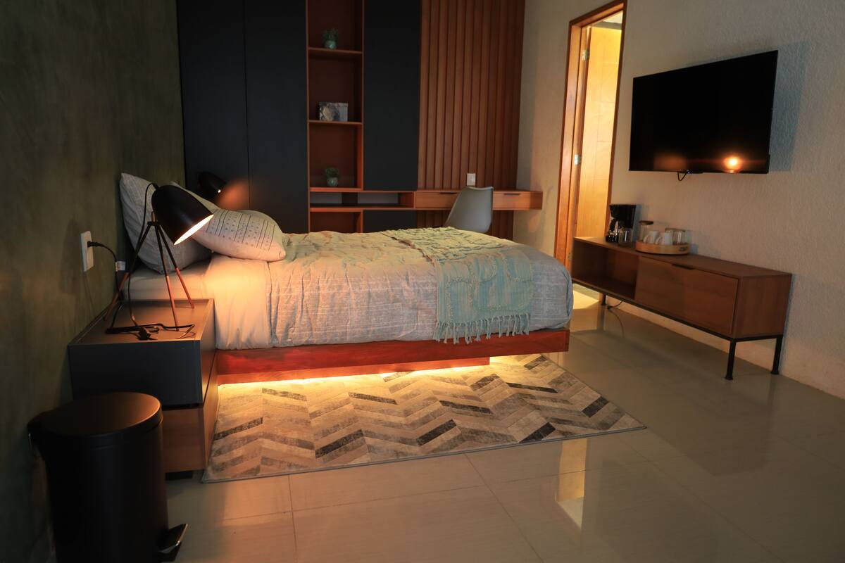 Quarto moderno com cama, mesa de cabeceira, luminária, TV e escrivaninha de madeira.