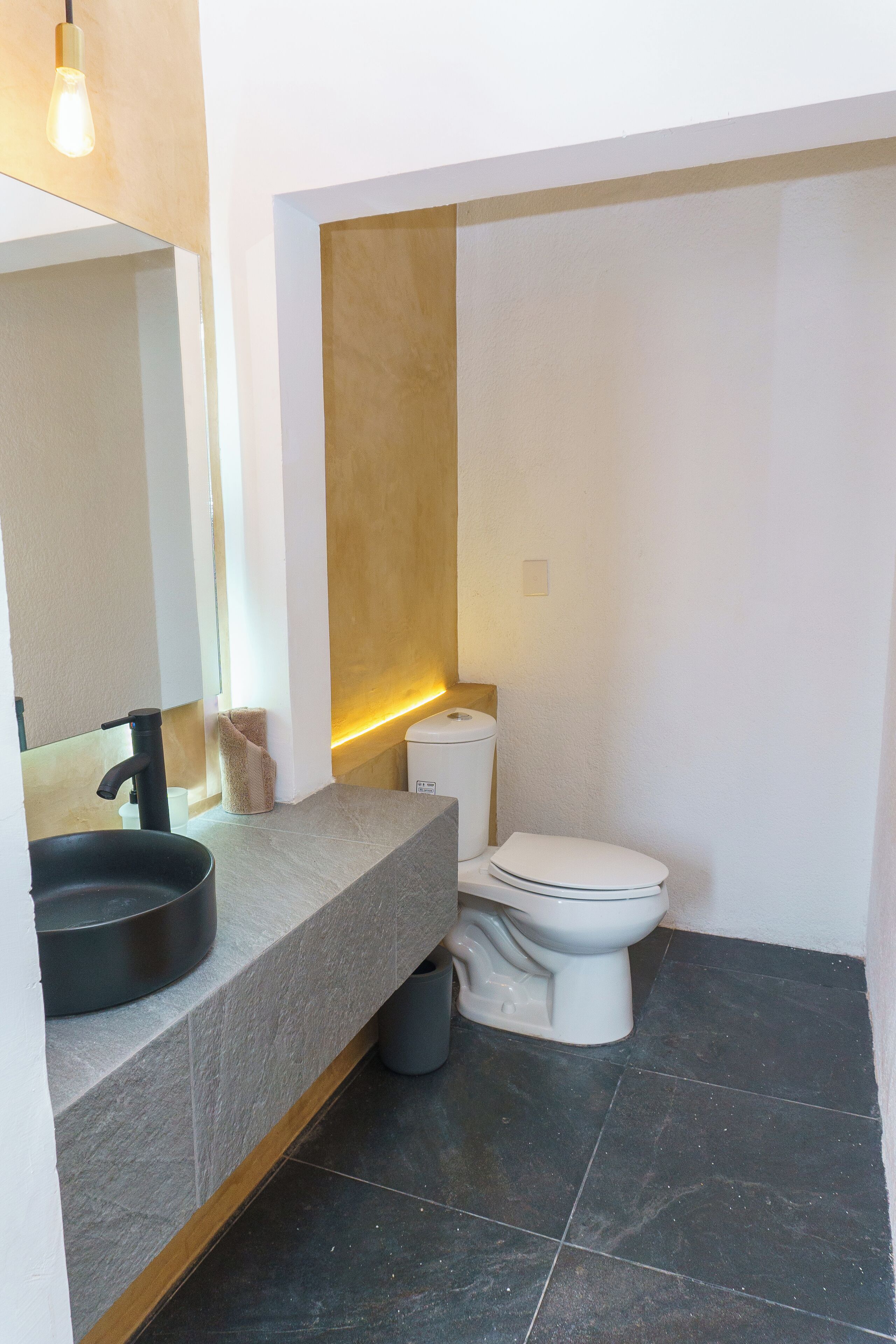 A modern bathroom with a grey countertop, a black sink, a white toilet, and a mirror.