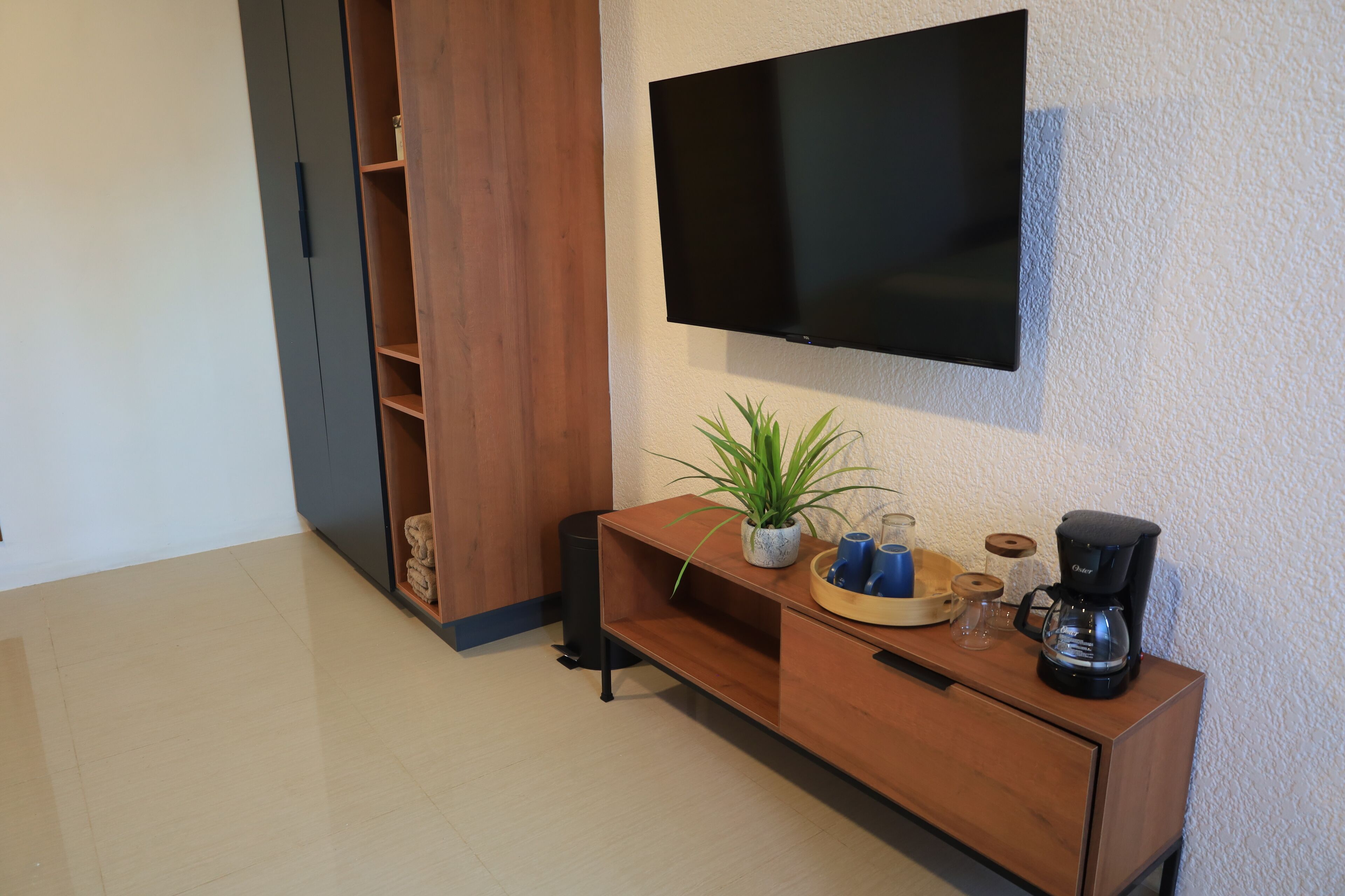 A modern living room with a flat-screen TV mounted on the wall, a wooden cabinet with a plant and a coffee maker, and a small trash bin.