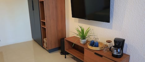 A modern living room with a flat-screen TV mounted on the wall, a wooden cabinet with a plant and a coffee maker, and a small trash bin.