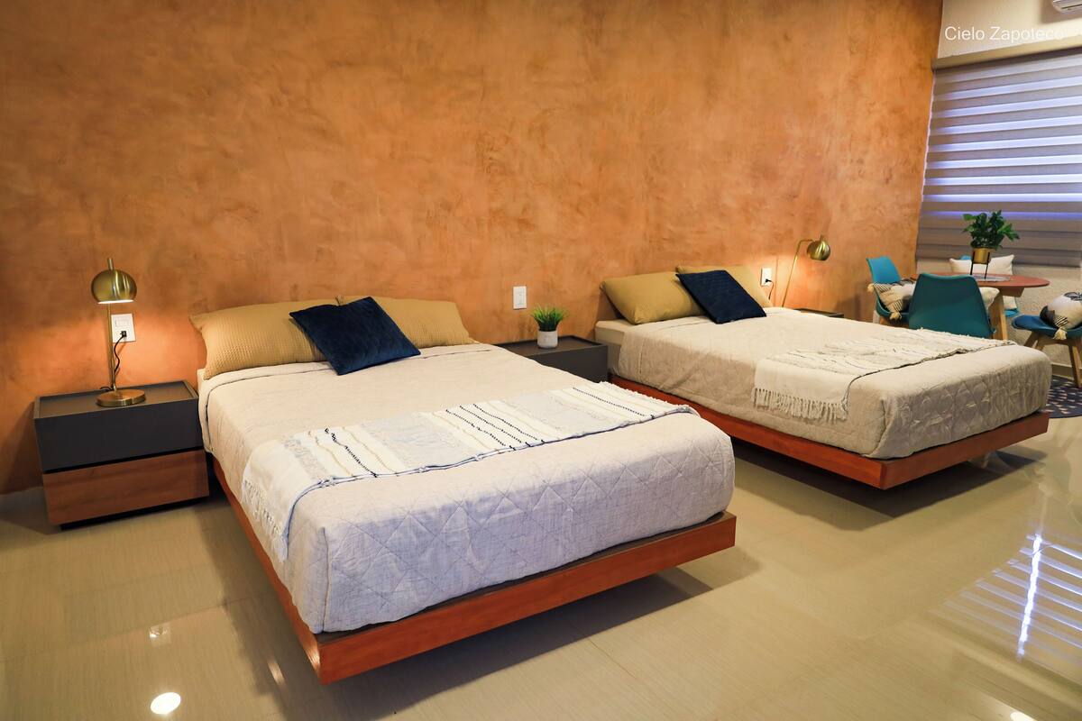 Quarto moderno com duas camas, mesas de cabeceira, uma pequena mesa e um vaso de planta.