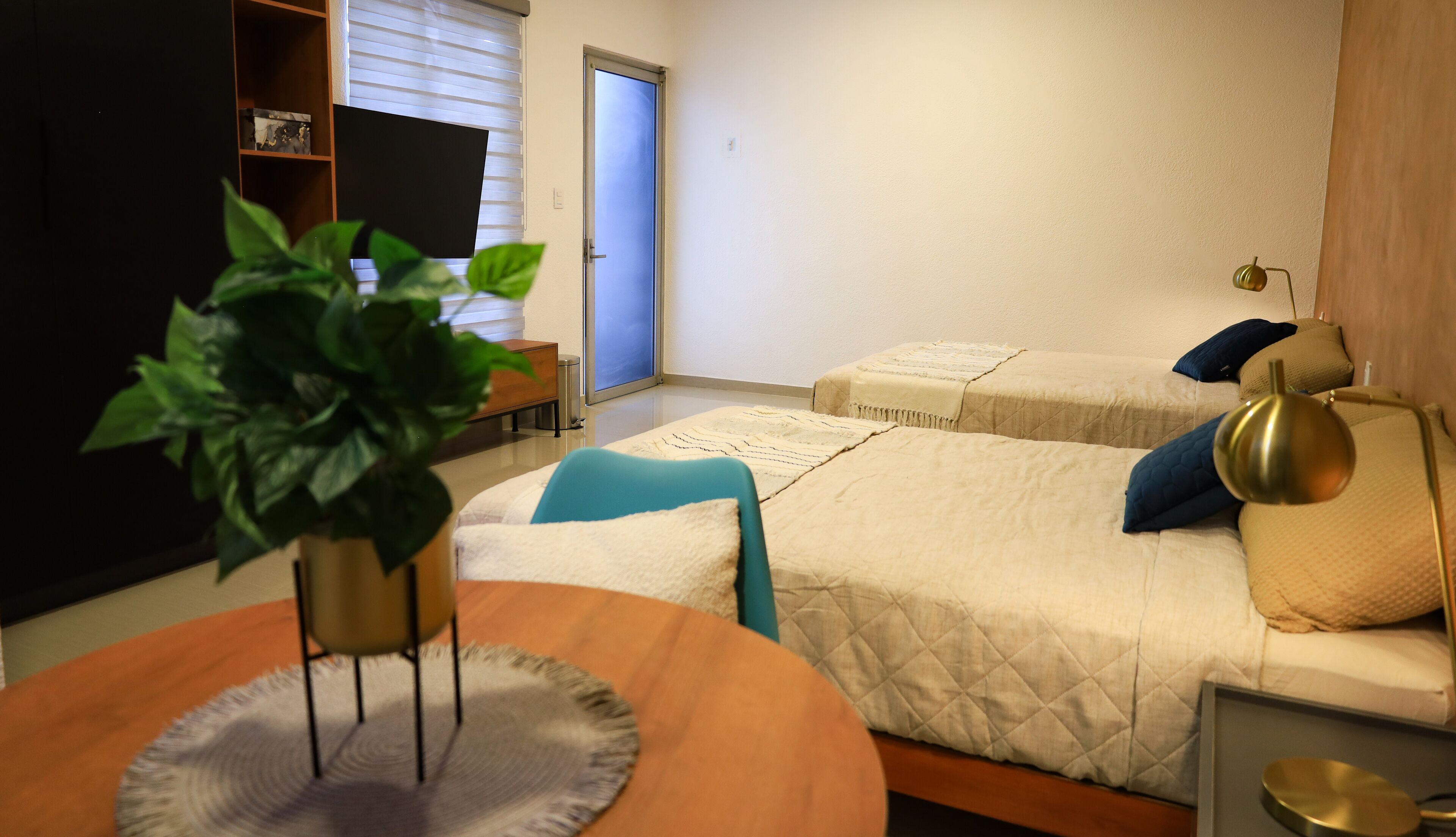 A hotel room with two beds, a desk with a chair, a small table with a plant, and a window with blinds.