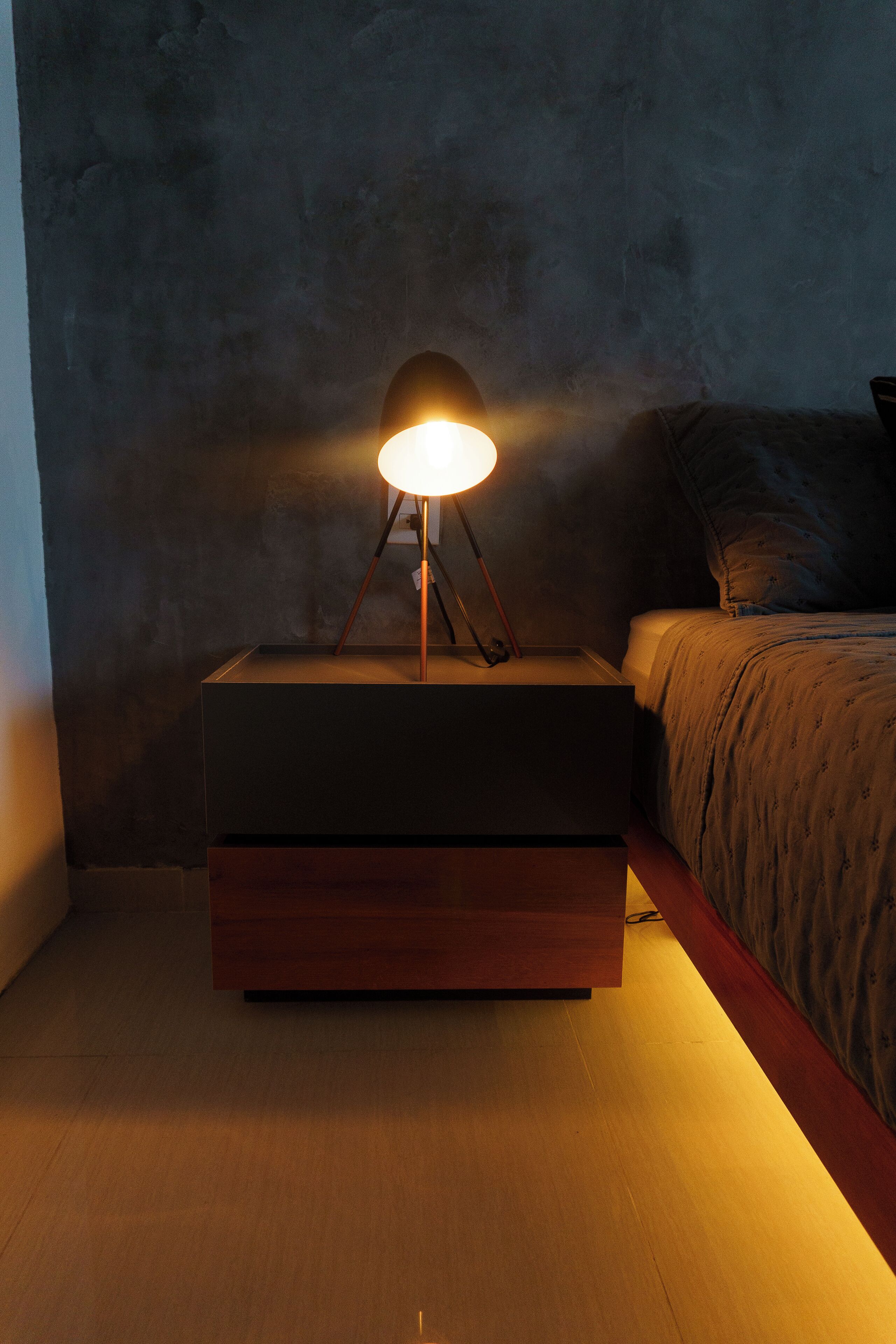 Una mesita de noche con una lámpara, una cama con colcha estampada y una pared con textura.