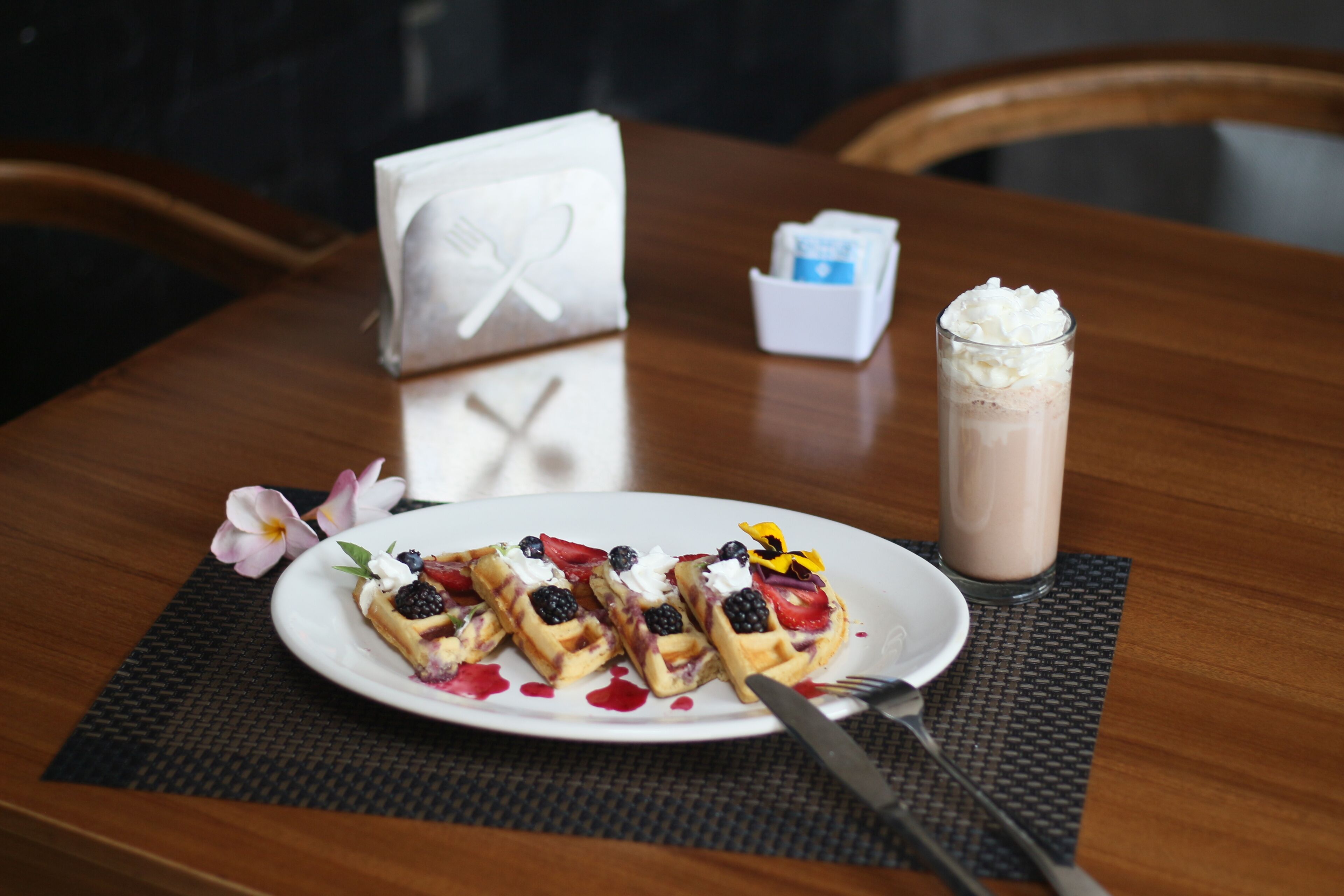 A plate of waffles with berries and whipped cream, a glass of chocolate milk, and a napkin with a fork on a wooden table.