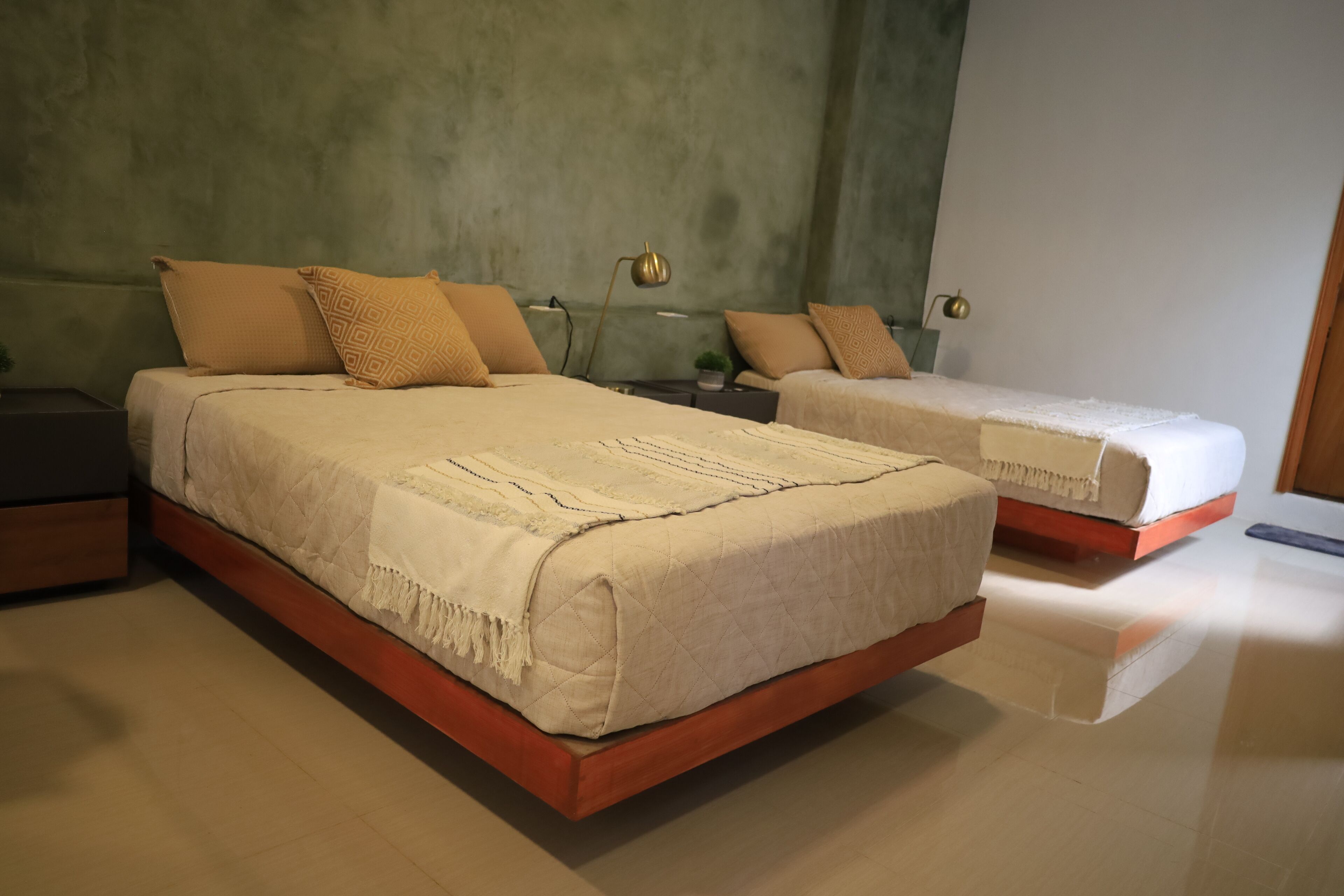 A bedroom with two beds, each with a wooden frame and beige bedding. There are bedside tables with lamps and a small plant on one of them.