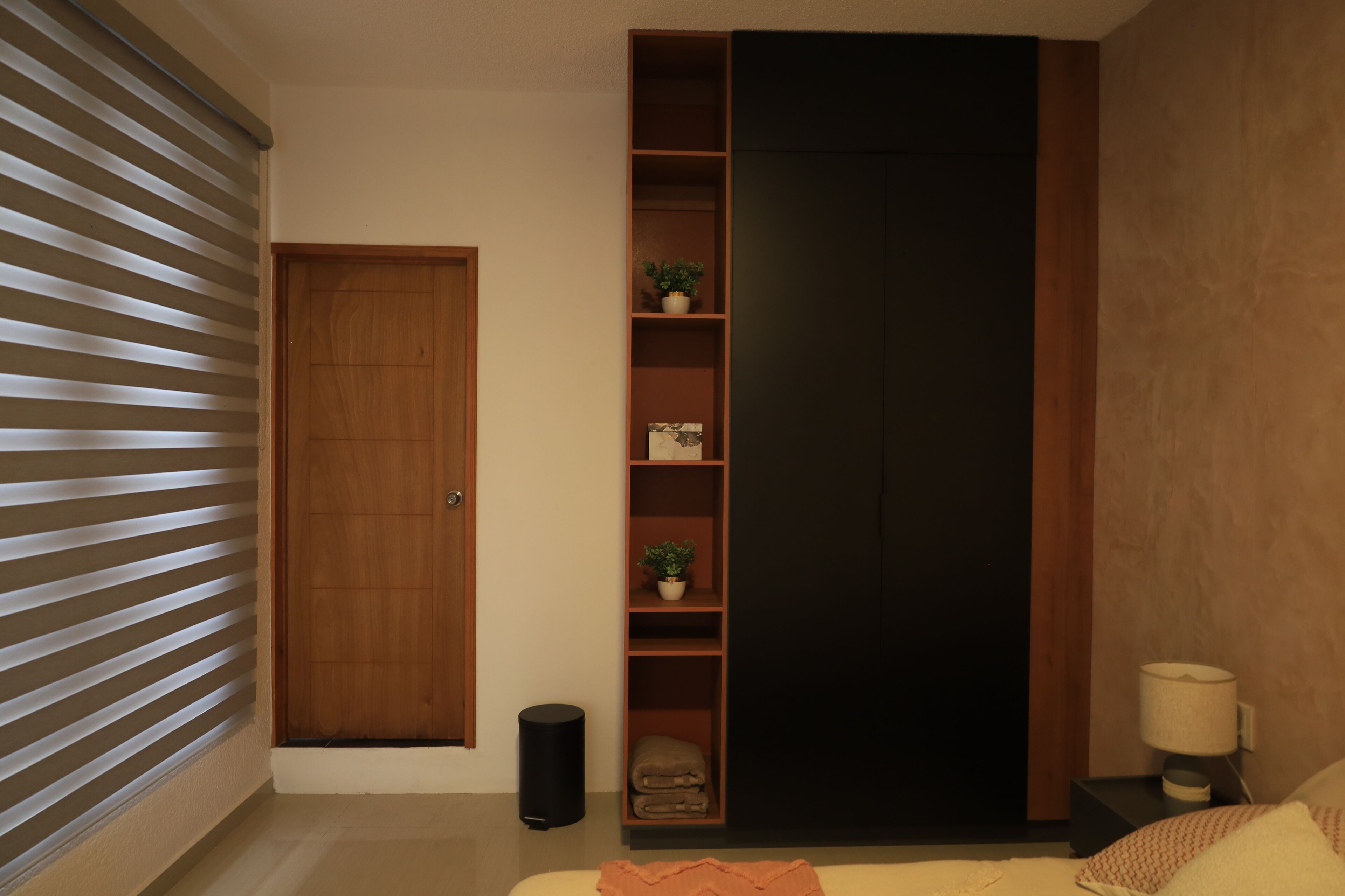 A bedroom with a wardrobe, a bedside table with a lamp, and a potted plant on the shelf.
