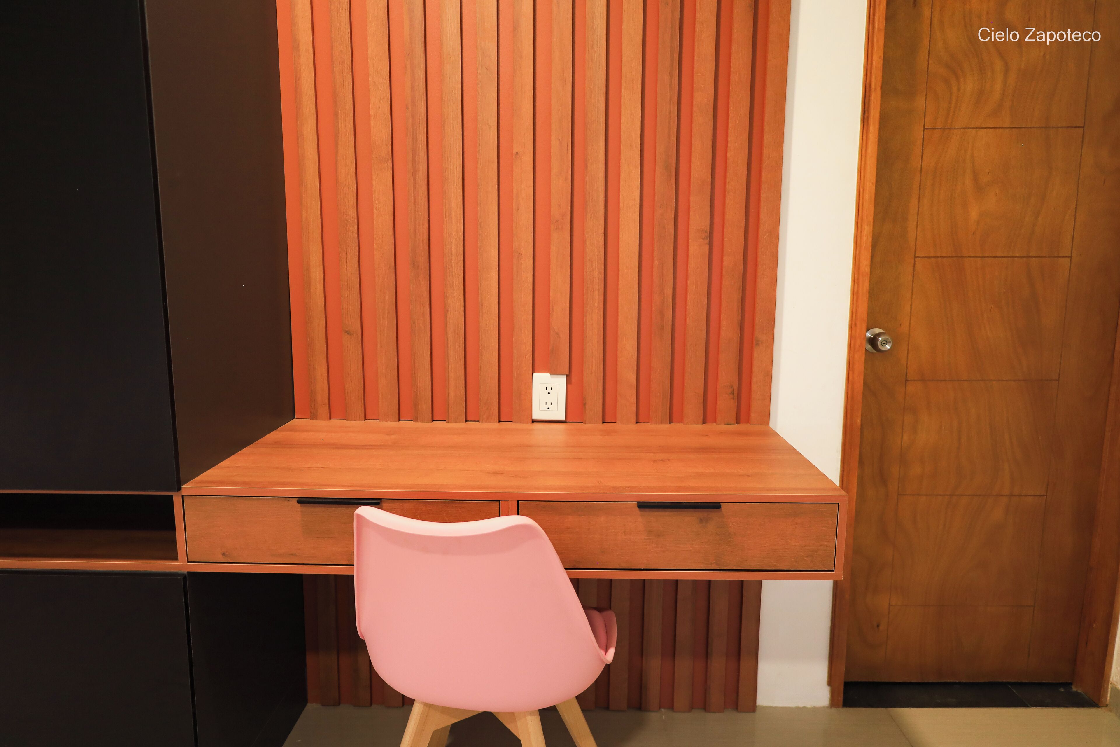 A wooden desk with a pink chair in front, a wooden door to the right, and a wall with vertical wooden slats.