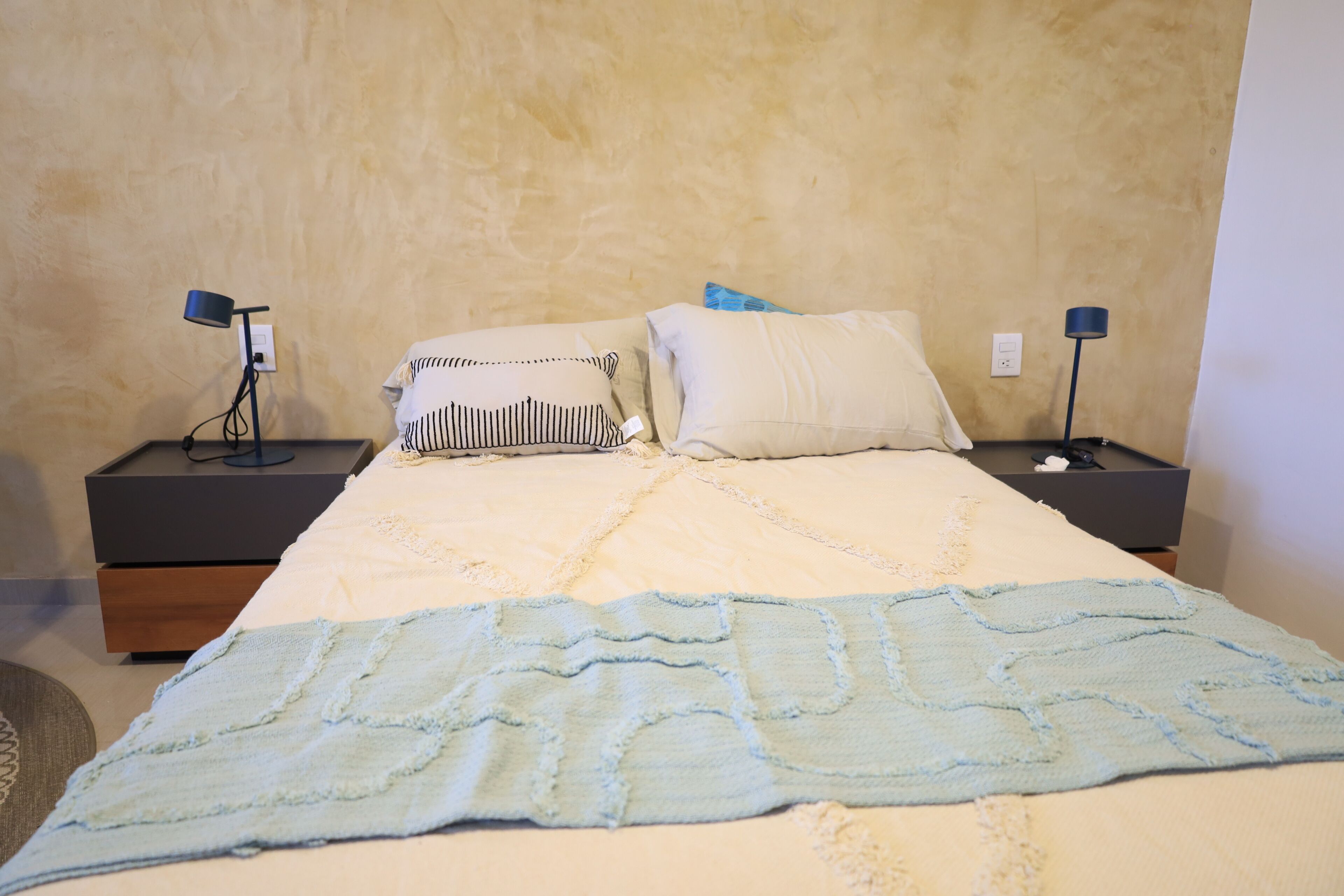 Un letto rifatto con cura, una coperta blu, due comodini con lampade e un muro con texture.