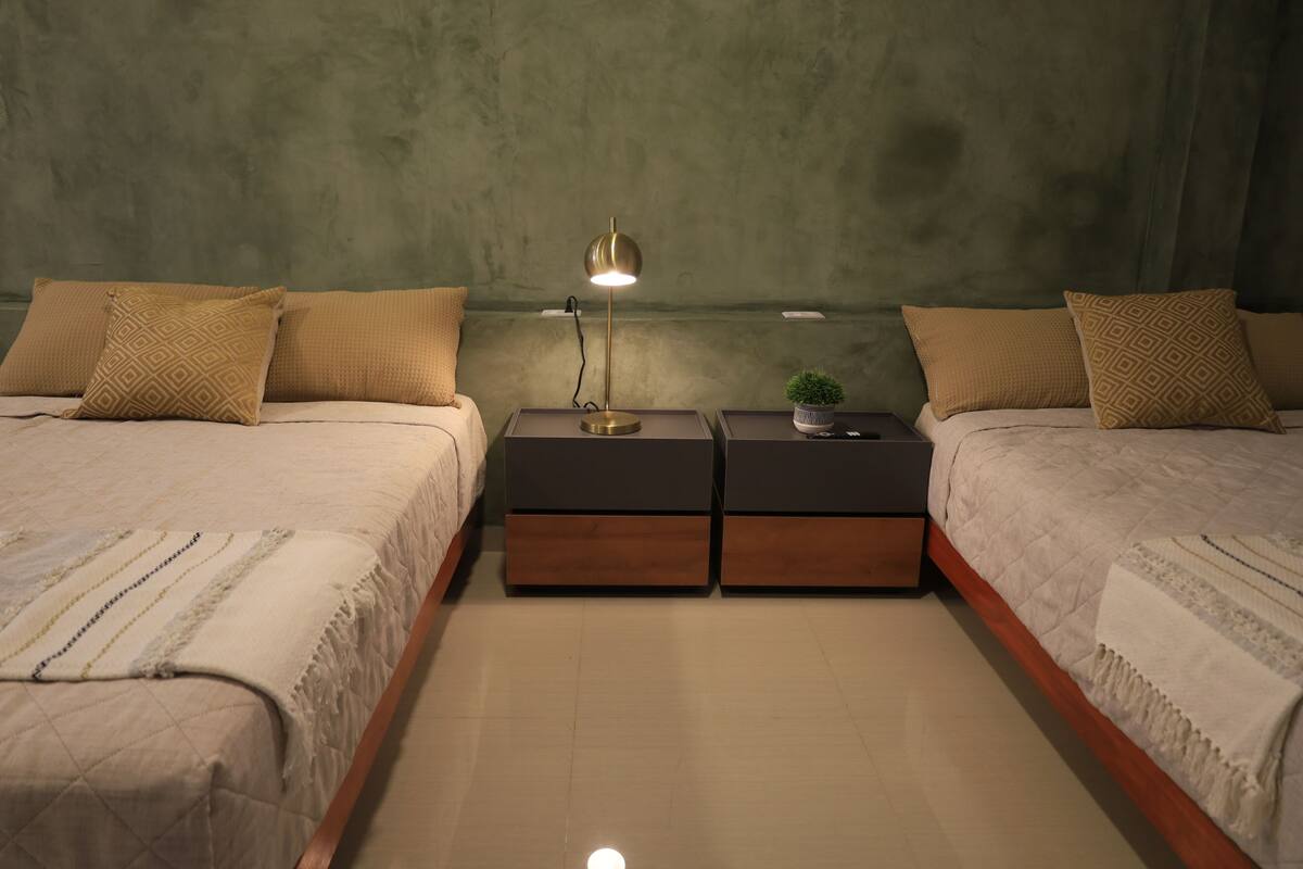Quarto com duas camas, mesas de cabeceira, uma luminária e um vaso pequeno com planta.