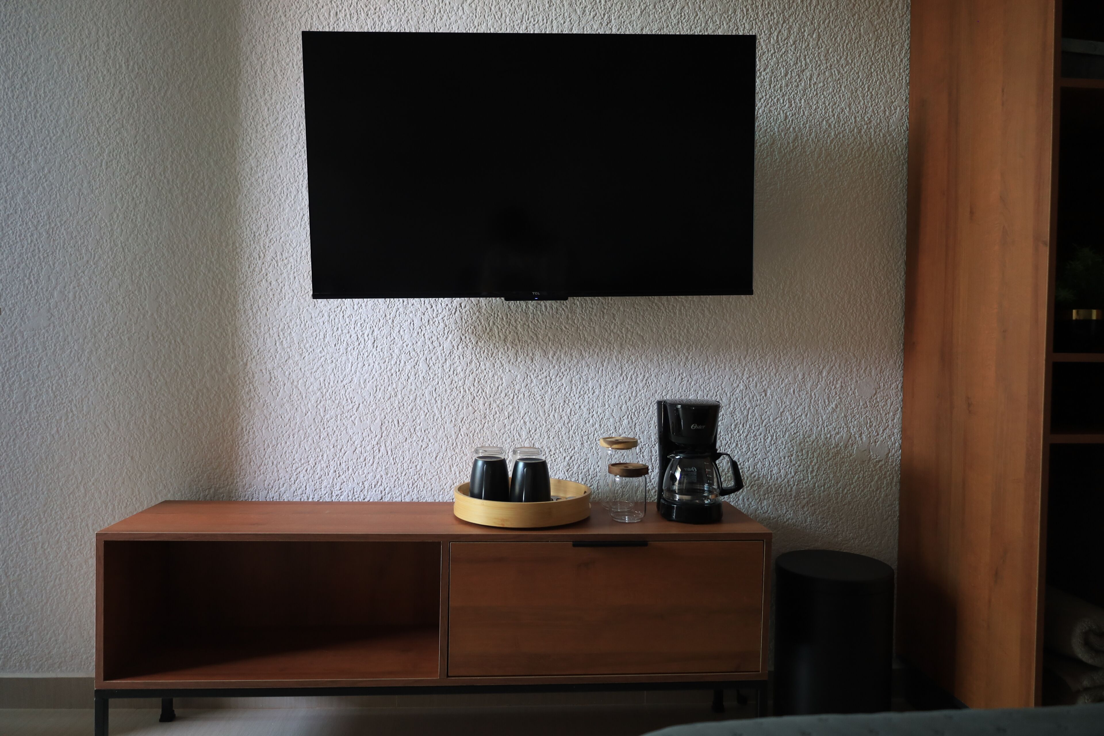Un salon moderne avec un téléviseur à écran plat fixé au mur, une table basse en bois sur laquelle se trouvent une cafetière et des verres, et un canapé en arrière-plan.
