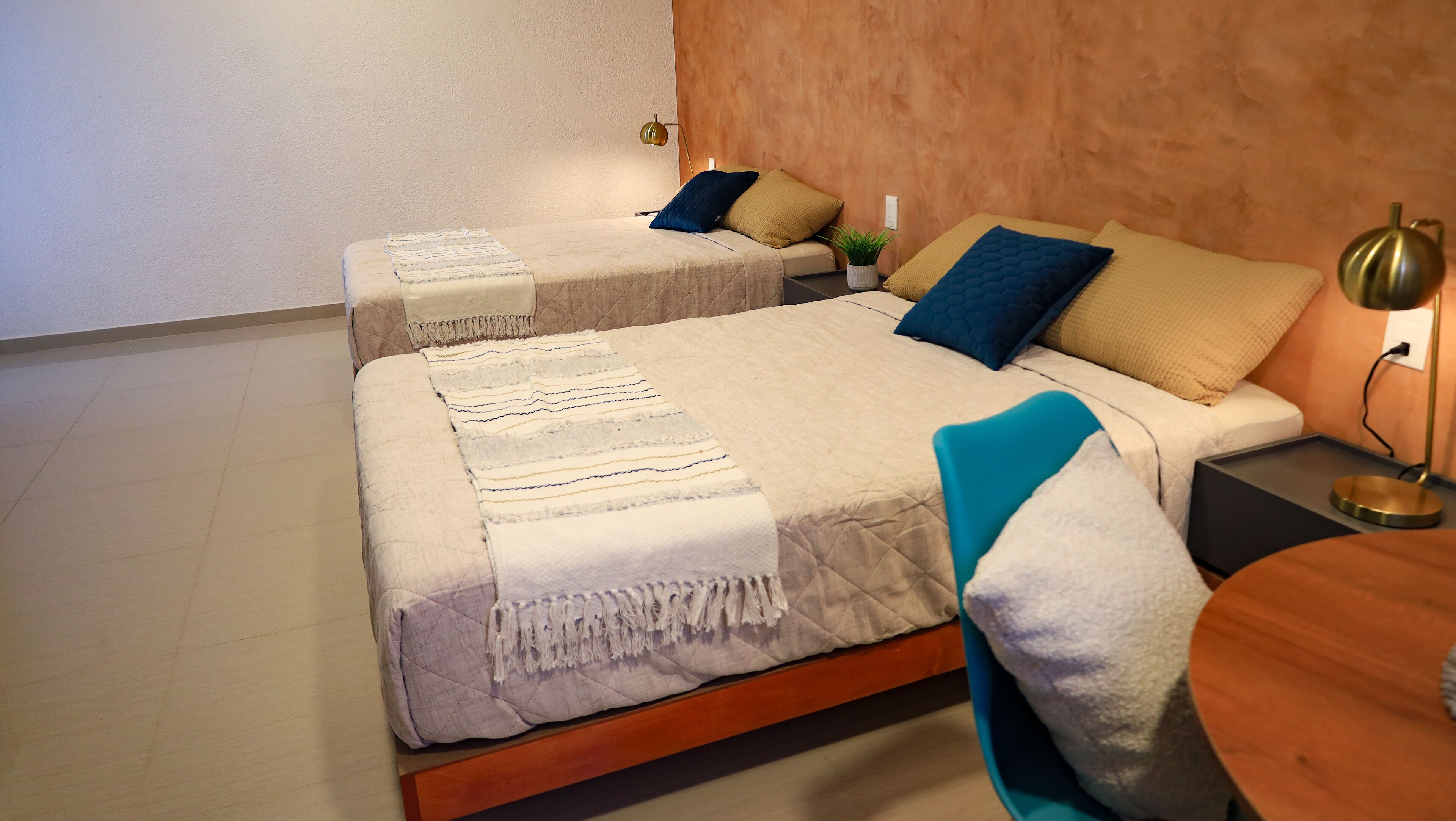 Een hotelkamer met twee bedden, elk met een andere soort beddengoed en kussens. Er staat een klein tafeltje met een lamp en een potplant aan de linkerkant van de kamer.