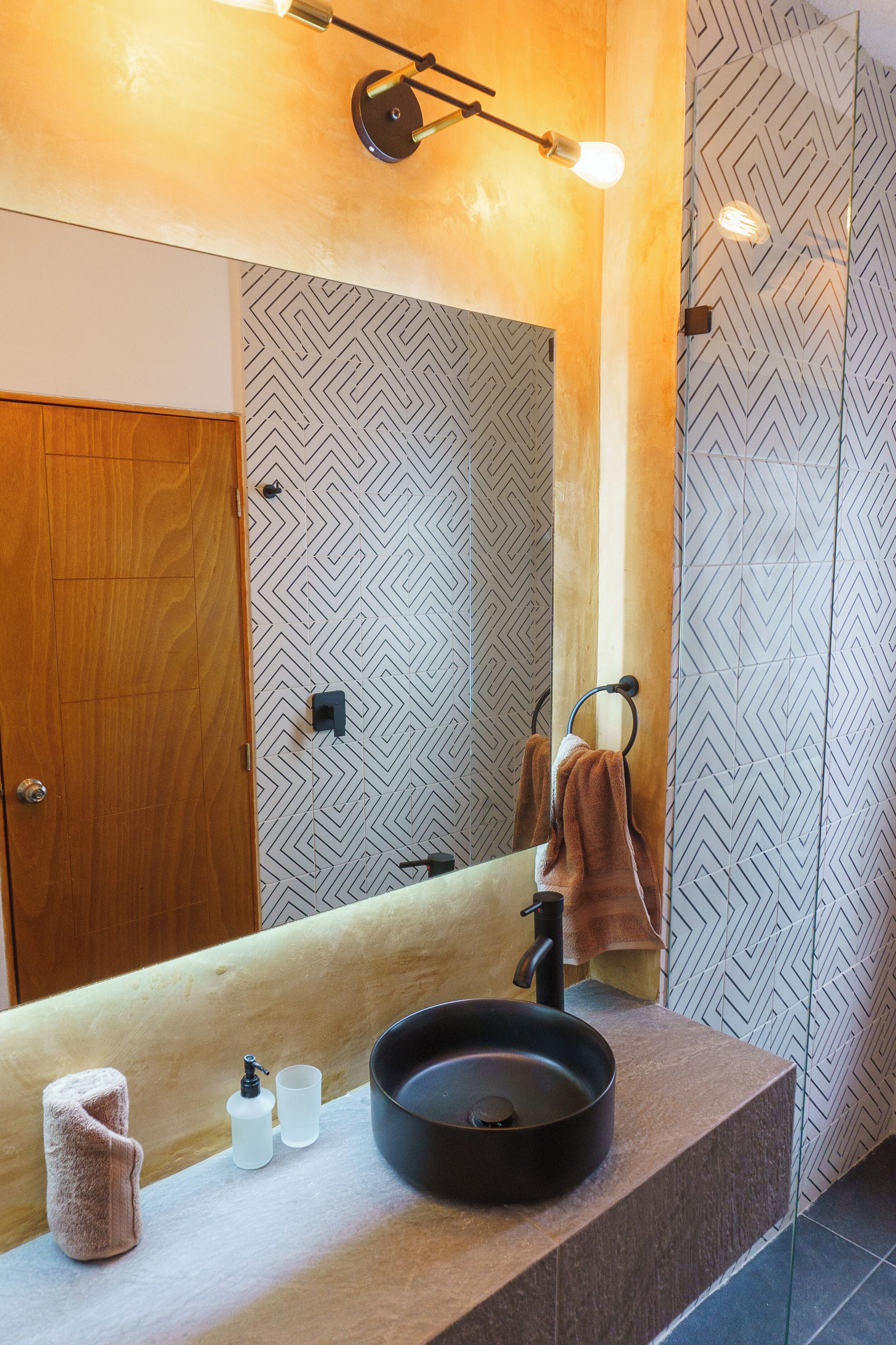 Une salle de bain avec une porte en bois, un miroir, un lavabo noir, une serviette et un distributeur de savon.