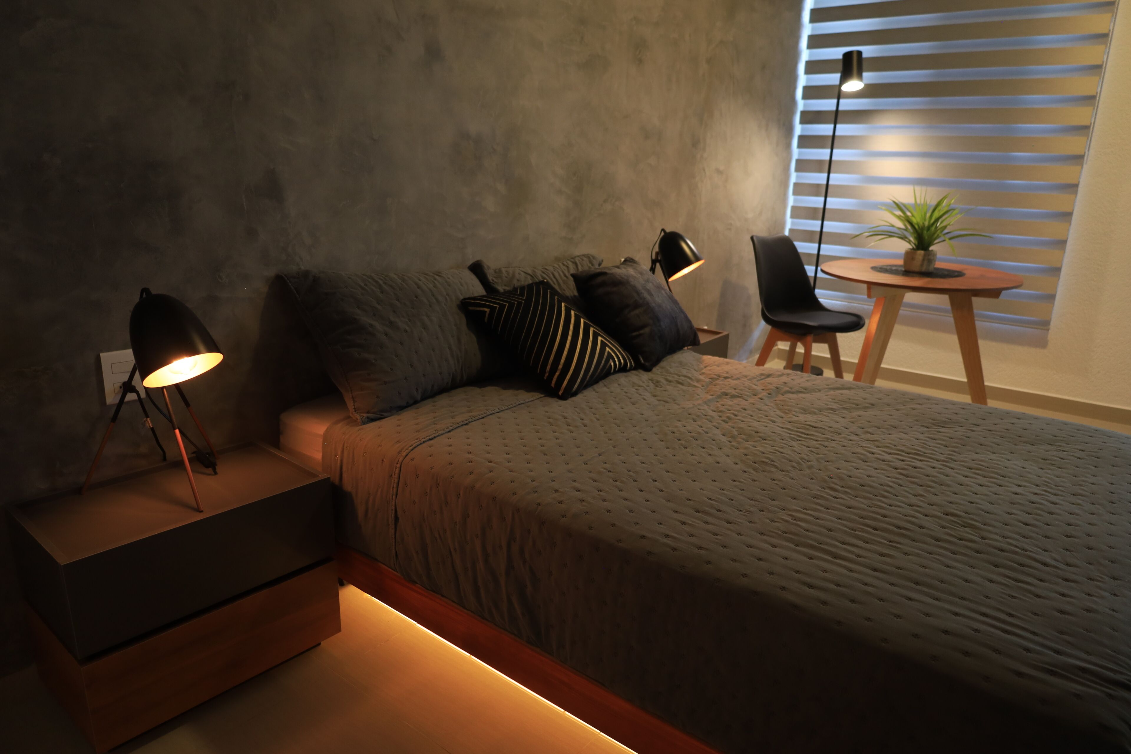 Una camera da letto moderna con un letto, comodini, una sedia, un tavolino con una pianta e una finestra con persiane.