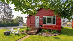 A small red house with a white door, a mailbox, and a front yard with grass and a white chair. - Cozy Corner - East end home (Ely)