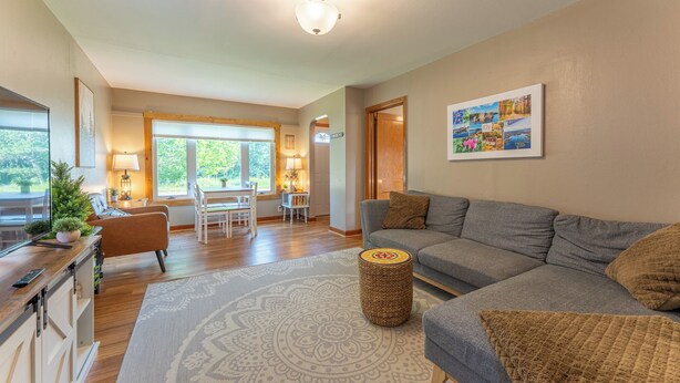A spacious living room with a grey sofa, a coffee table, and a dining area with a table and chairs. There is a large window providing natural light and a view of greenery outside. - Cozy Corner - East end home (Ely)