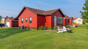 A red single-story house with a front porch, a garage, and a well-maintained lawn. - Cozy Corner - East end home (Ely)