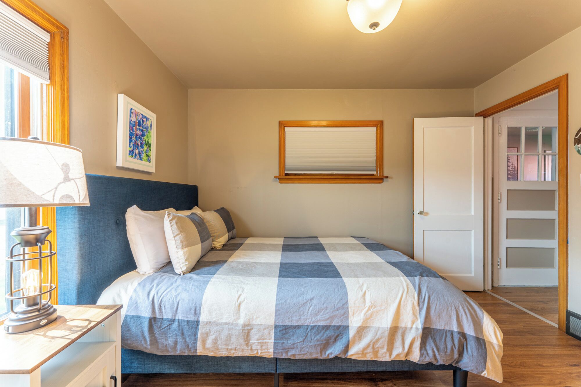 Un dormitorio con cama, mesita de noche, lámpara, ventana con persianas, puerta y un cuadro en la pared.
