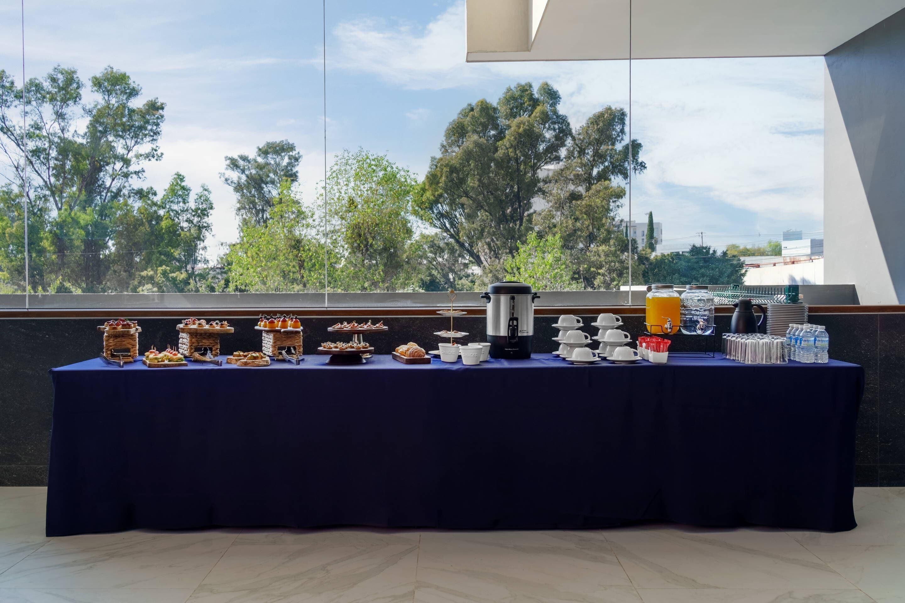 A buffet table with various food items and beverages, set against a backdrop of trees and a clear sky.