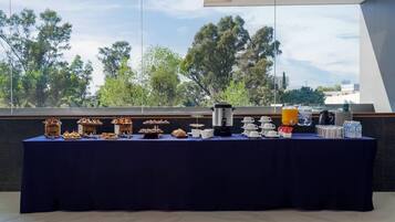Una mesa de buffet con diversos alimentos y bebidas, con árboles y un cielo despejado de fondo.