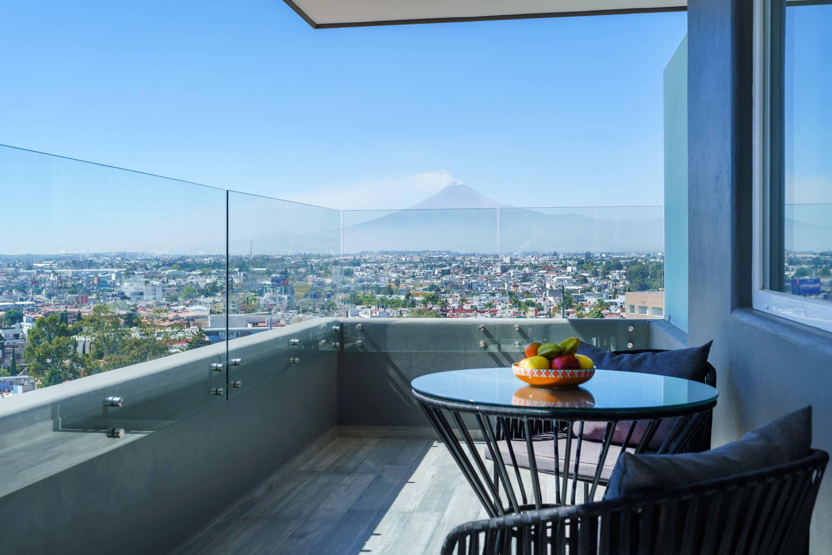 A balcony with a glass railing, a round table with chairs, and a bowl of fruit.
