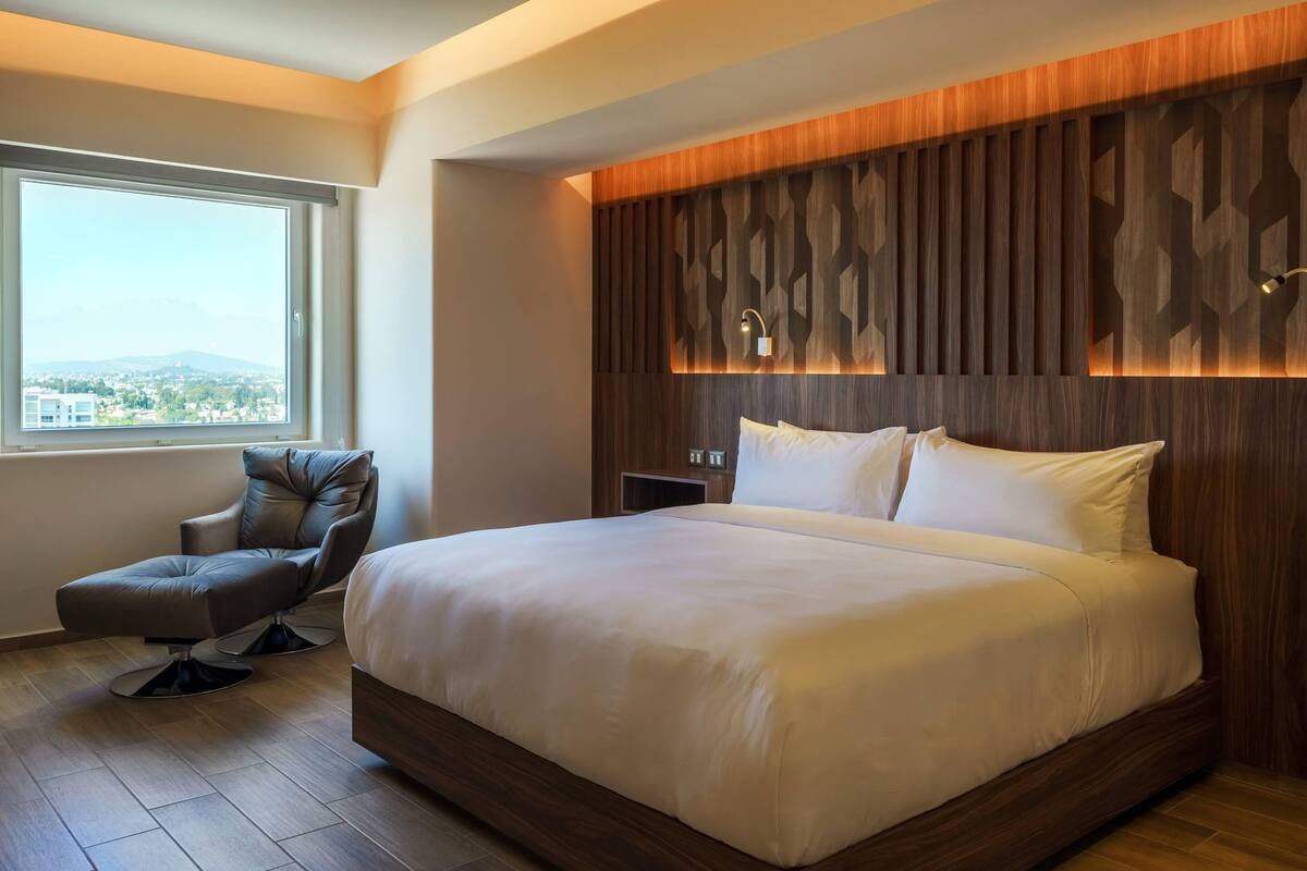 A modern hotel room with a large bed, a sitting area with a chair, a nightstand, and a view through a large window.