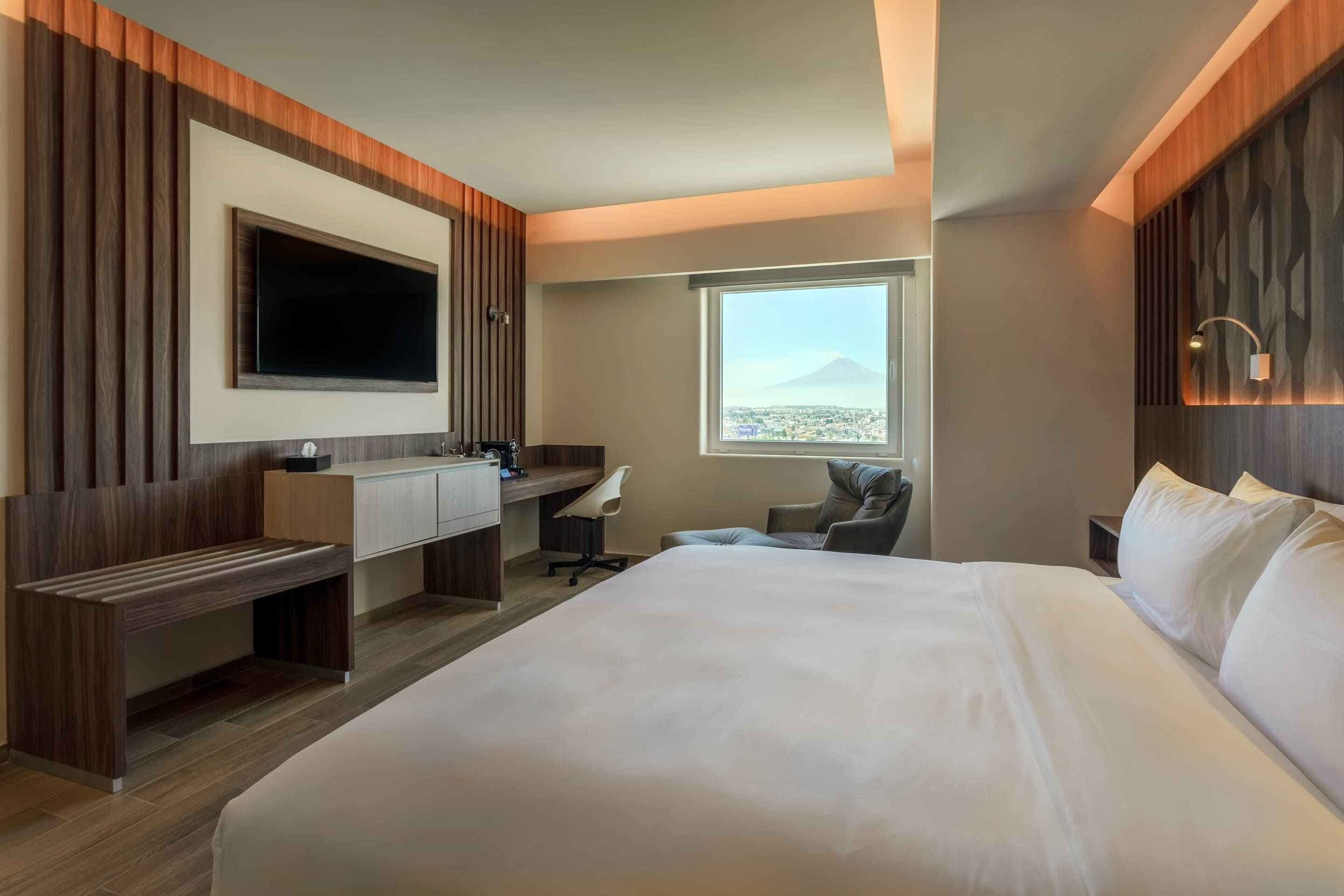 A hotel room with a large bed, a desk with a chair, a TV mounted on the wall, and a window with a view of a mountain.