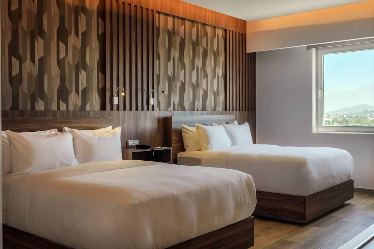 A hotel room with two beds, a wooden wall panel, a nightstand, and a window with a city view.