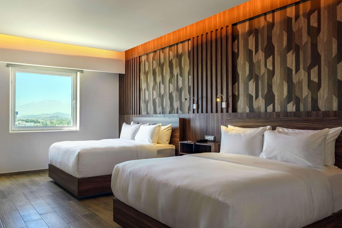 Habitación de hotel con dos camas, un ventanal grande y un panel decorativo en la pared.