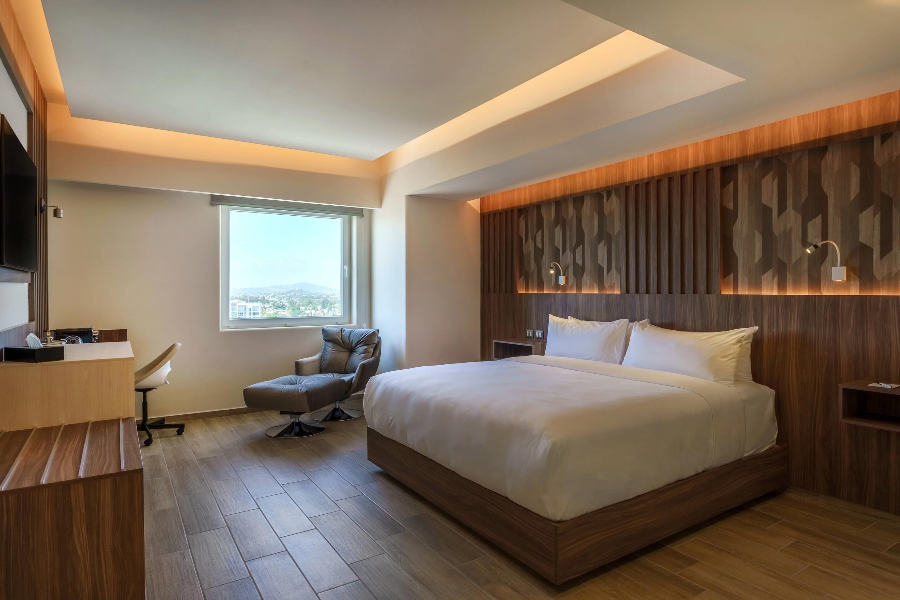 A modern hotel room with a large bed, a desk, a chair, and a view of the city through a window.
