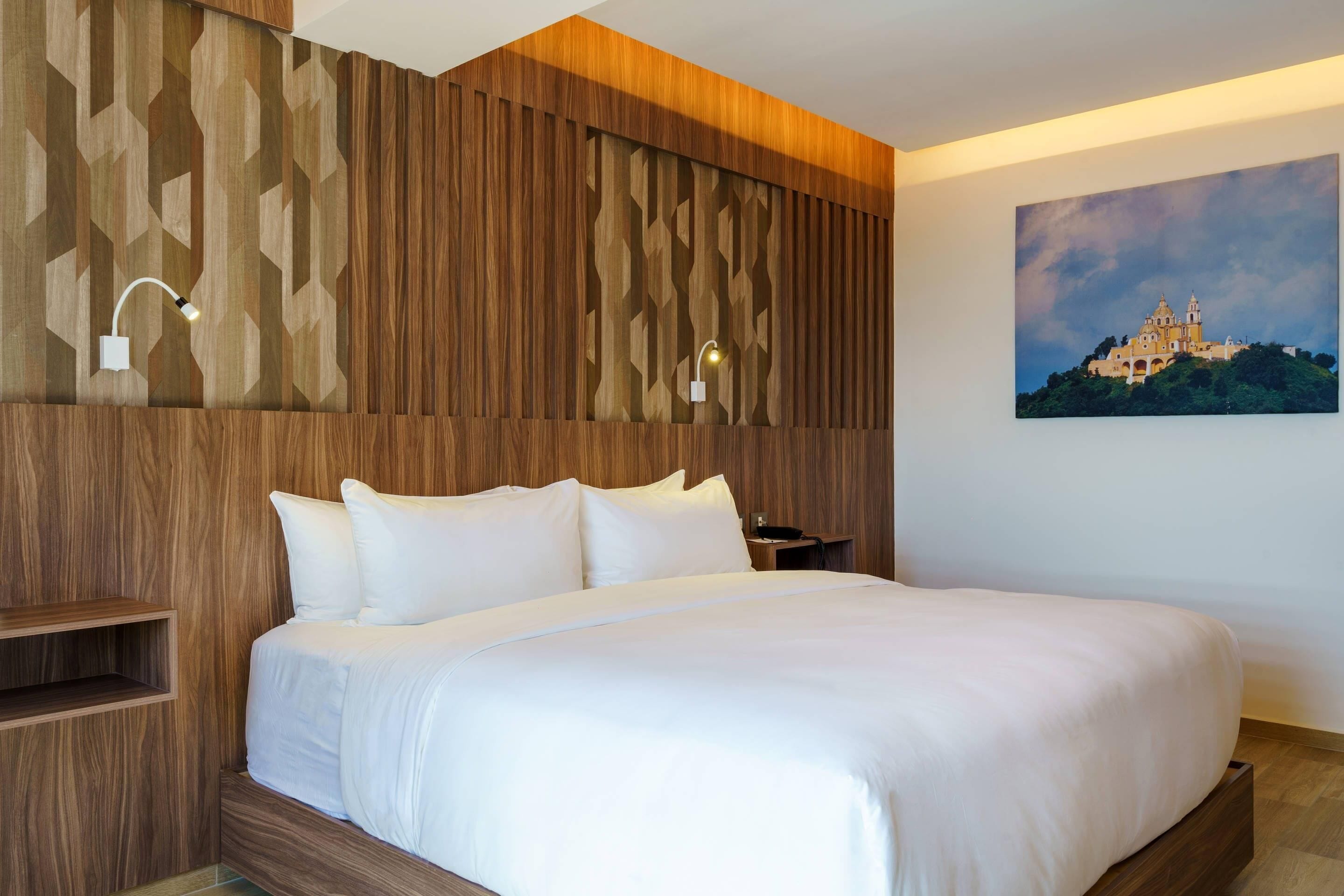 A hotel room with a wooden headboard, a bed with white linens, a nightstand, and a wall-mounted lamp.