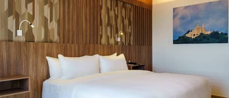 Una habitación de hotel con cabecera de madera, cama con ropa blanca, mesita de noche y lámpara de pared.