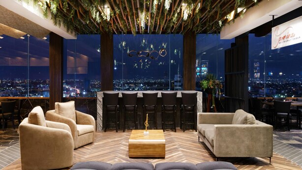 A modern lounge area with a city view, a bar with high stools, and plush seating. - Radisson Puebla Angelopolis (Puebla)