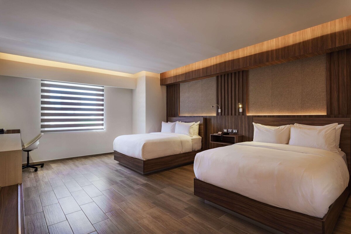 A hotel room with two beds, a desk, and a large window with blinds.