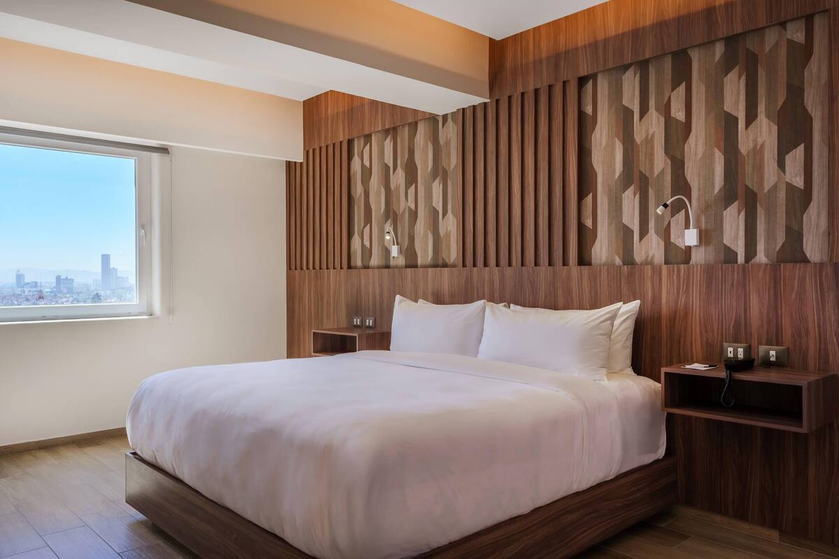 A modern hotel room with a large bed, wooden panel wall, bedside tables, and a city view.