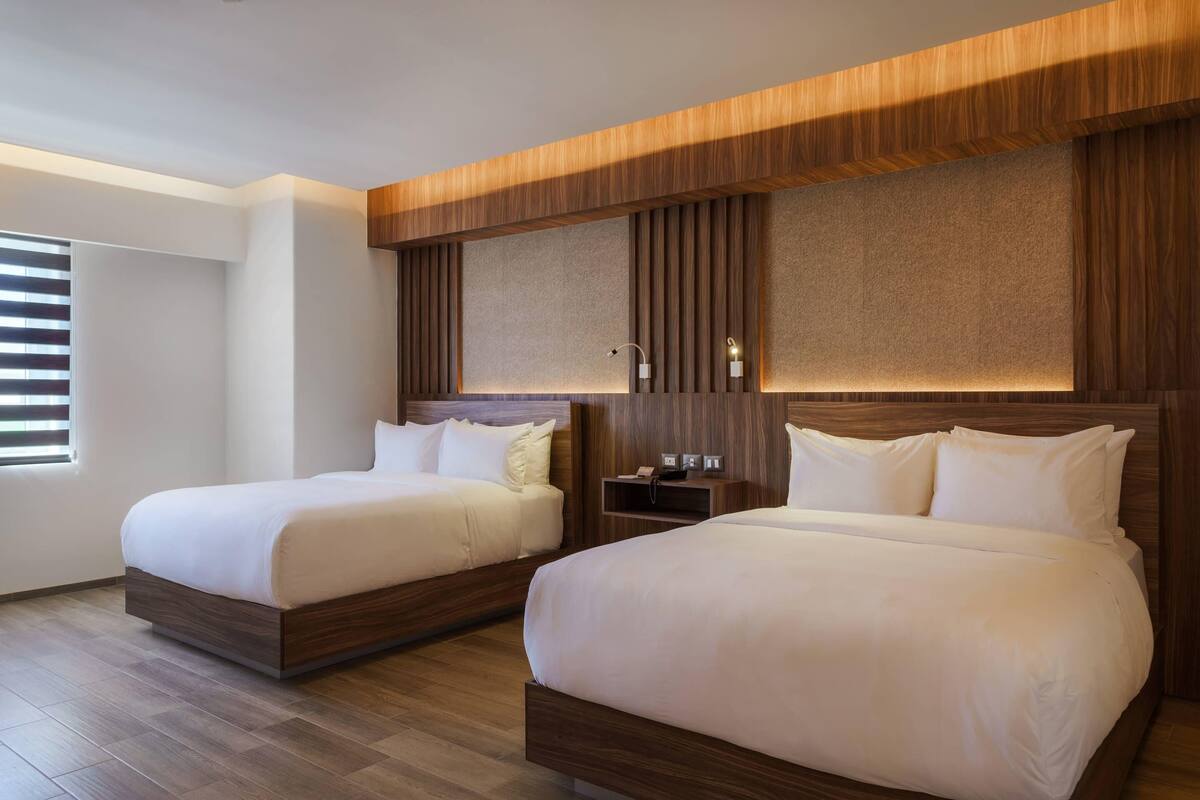 A hotel room with two beds, wooden headboards, a nightstand, and a window with blinds.