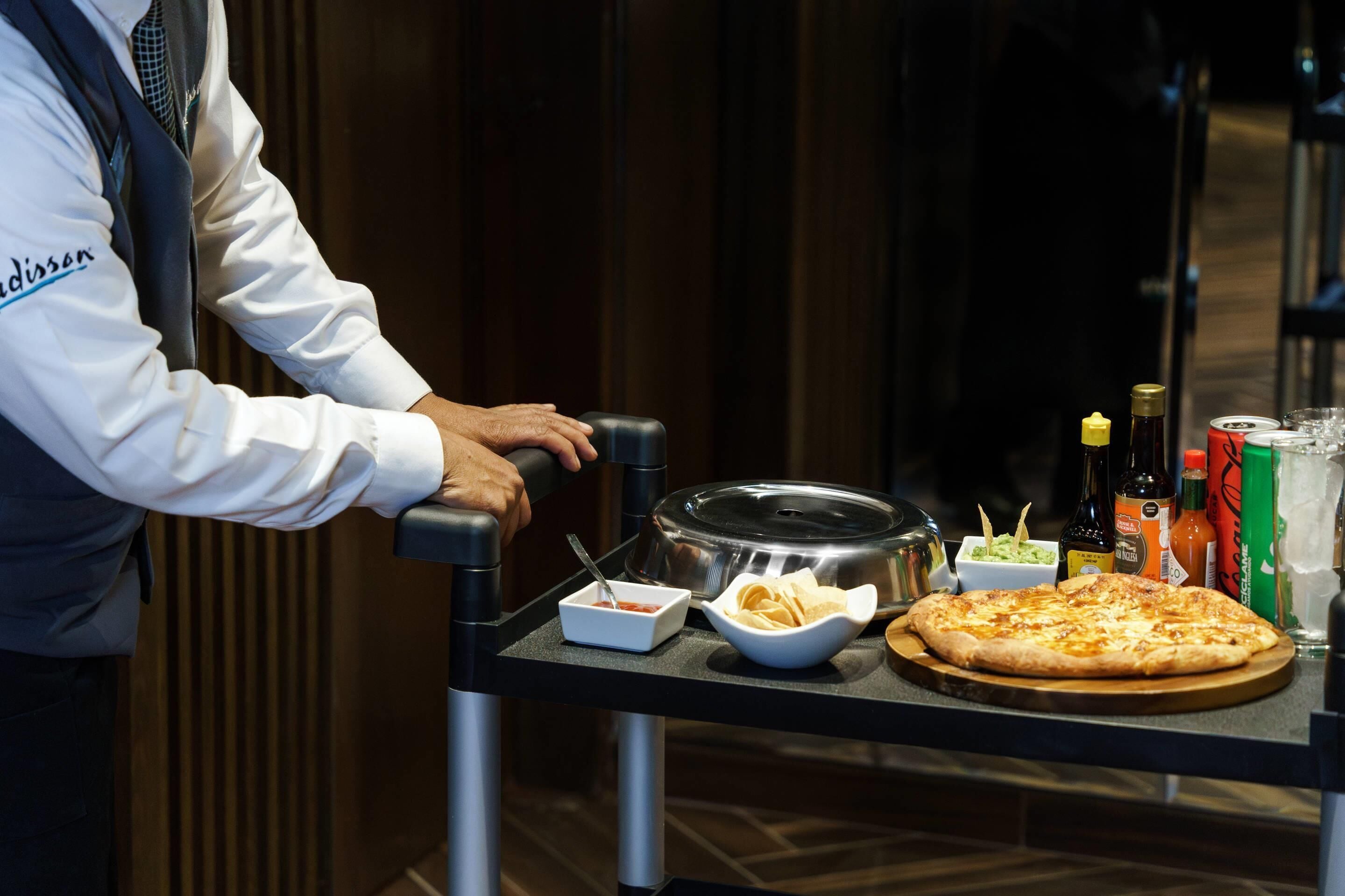 A person serving a pizza with various condiments and drinks on a tray.